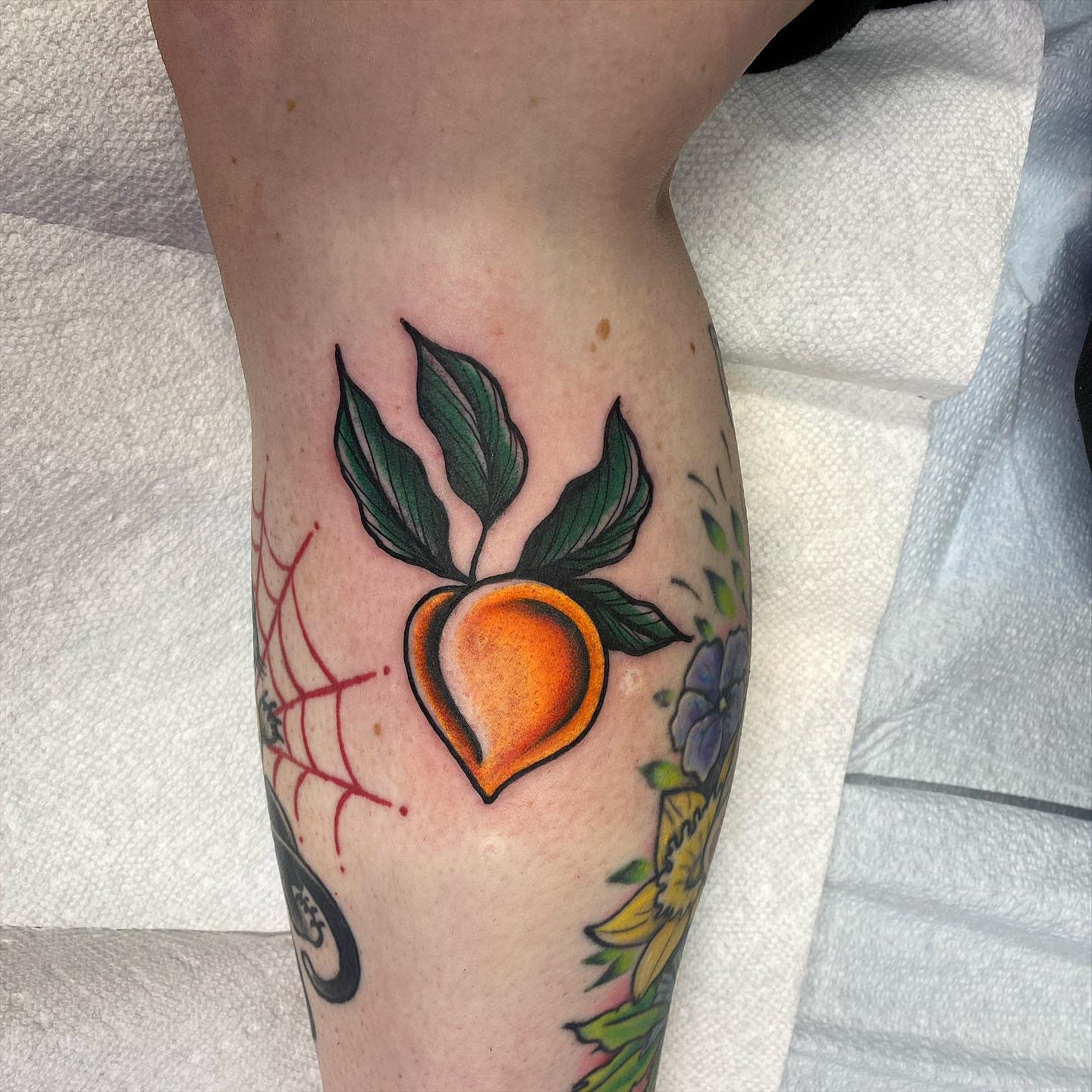A tattoo of a peach with leaves on a leg, surrounded by other colorful tattoos